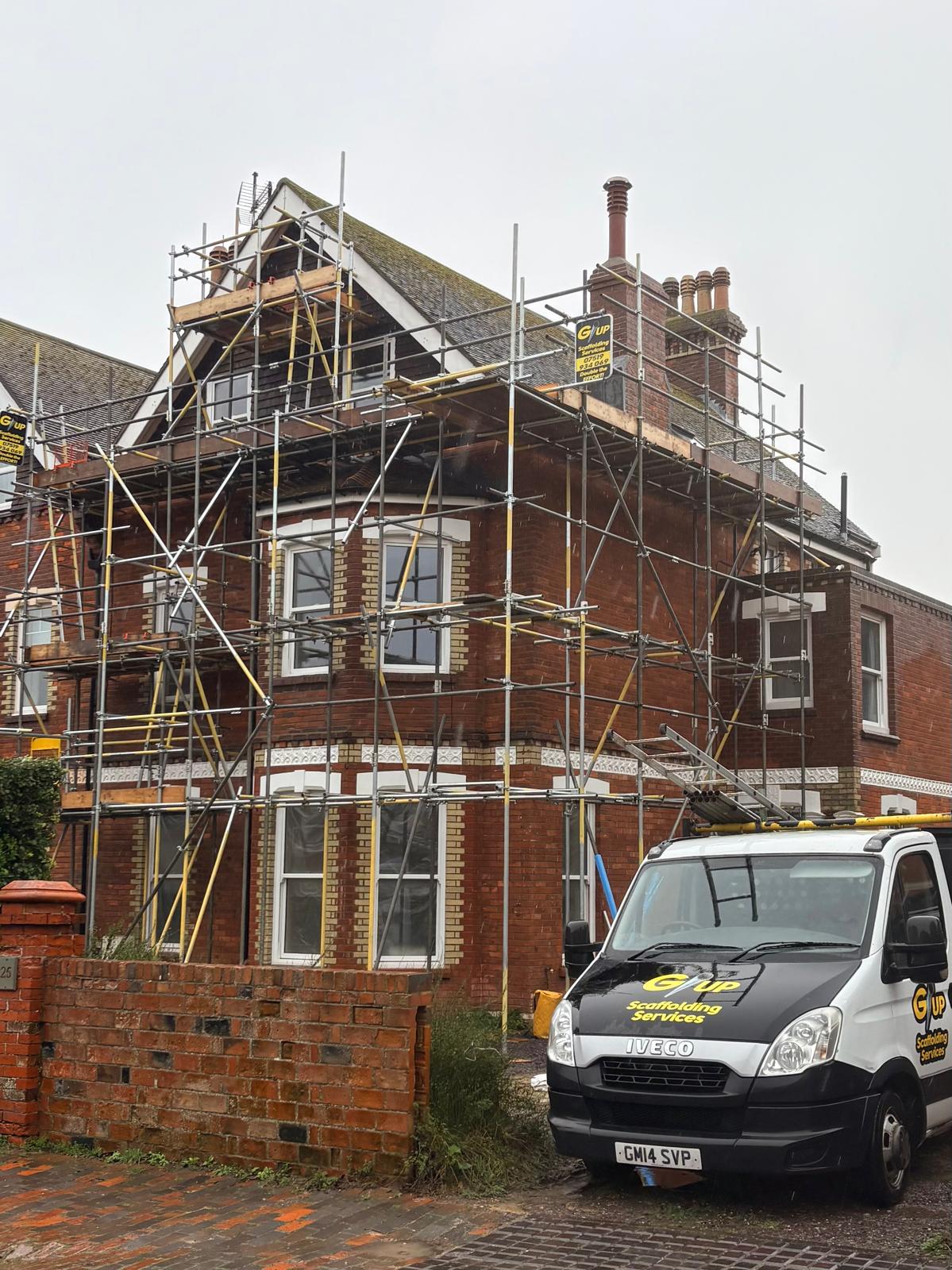 Scaffolding erected around a brick house for roofing works, with the G/UP van on site